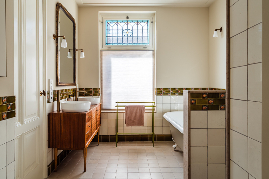 De badkamer met historische vloer- en wandtegels, glas-in-lood raam en bad op pootjes